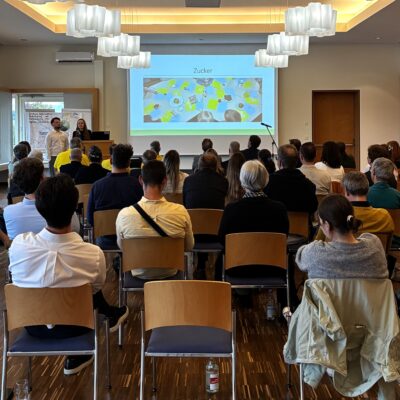 Eine Vertreterin und ein Vertreter des Kinderhauses St. Martin stehen am Podium und zeigen eine Präsentation zu ihrem Gewinnerprojekt, vor ihnen sitzen zahlreiche Gäste