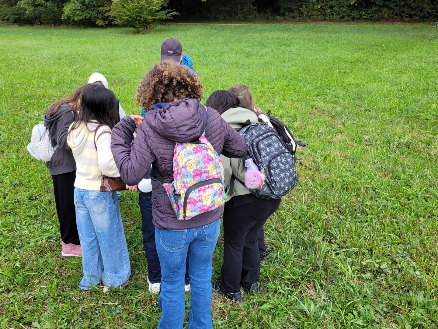 Acht Schülerinnen und Schüler stehen auf einer Wiese im Kreis und schauen auf etwas, das auf dem Bild nicht zu sehen ist.