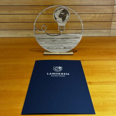 Runde Holztrophäe mit einer stilisierten Glühbirne, in die eine Weltkugel eingesetzt ist und mit Schriftzug Klimaschutzpreis 2026 Landkreis Konstanz